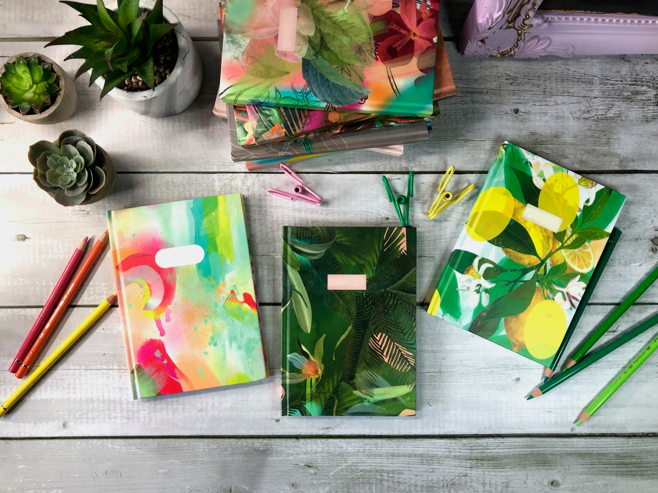 Colorful notebooks with tropical and abstract patterns on a wooden table, surrounded by pencils, pens, and potted plants.