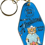 The Hank's Honkitonk Hideout Keyring Blue by Hôtel Dodo features a cat cowboy and rustic accents, with "Hotel Dog Resort" and "Hank's Honkitonk Hideout" text—a fun bag charm to add log cabin flair to your accessories.
