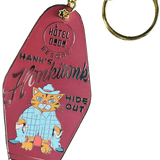 The Hank's Honkitonk Hideout Keyring by Hôtel Dodo features a vintage pink motel key tag design with a cartoon cat in a blue outfit, capturing the charm of an enamel tag reminiscent of a secluded rustic cabin.
