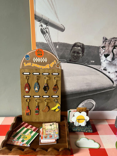 Two motel-style keychains with cartoon cats and text, one red and one blue, plus a small shoe-shaped charm, are on a wooden surface.