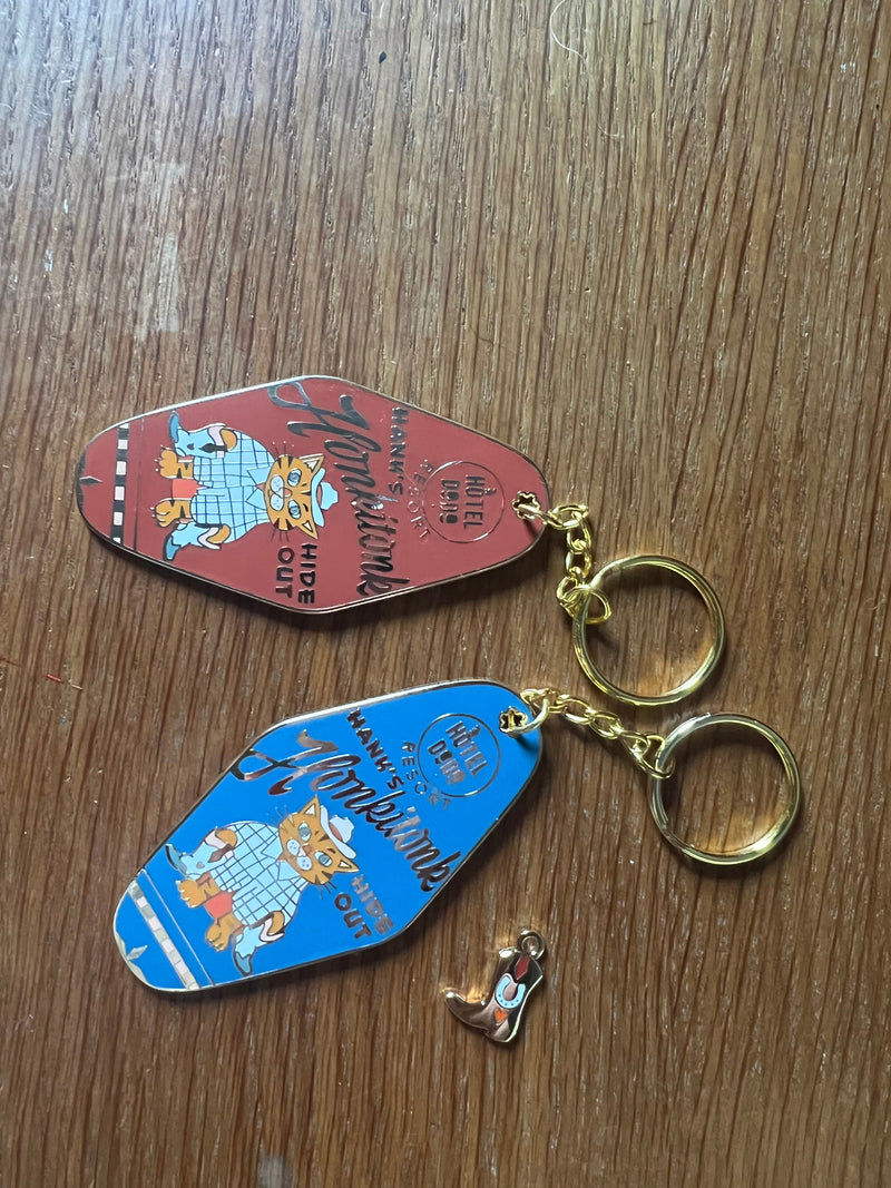 Two motel-style keychains with cartoon cats and text, one red and one blue, plus a small shoe-shaped charm, are on a wooden surface.