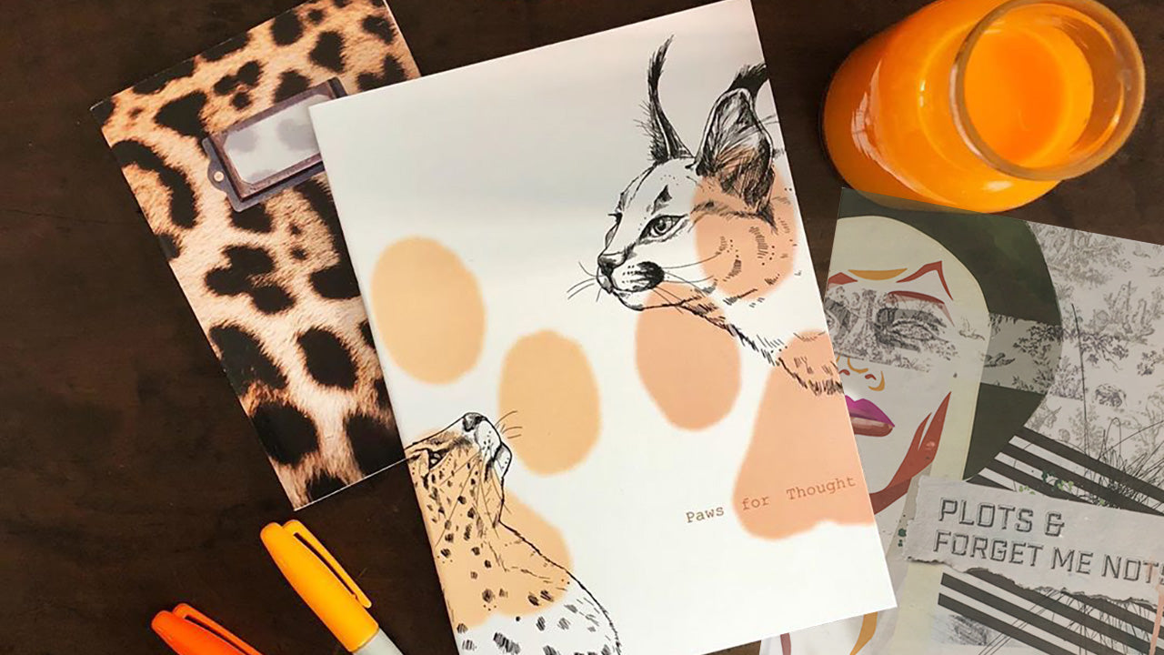 A desk with three notebooks—one with a leopard print cover, one with illustrated wild cats labeled "Paws for Thought," and one with a face and text, plus markers and a glass of orange juice.