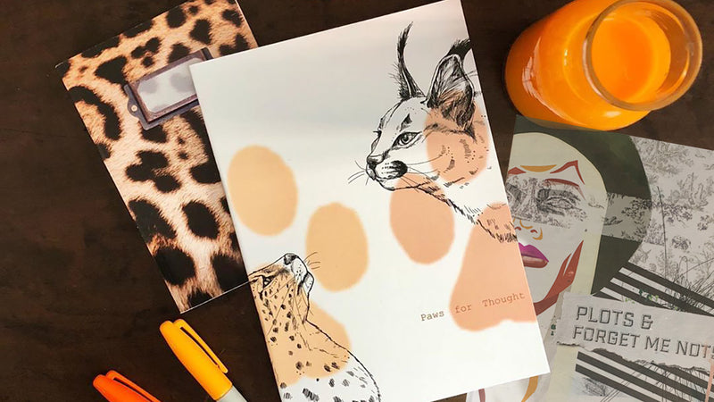 A desk with three notebooks—one with a leopard print cover, one with illustrated wild cats labeled "Paws for Thought," and one with a face and text, plus markers and a glass of orange juice.