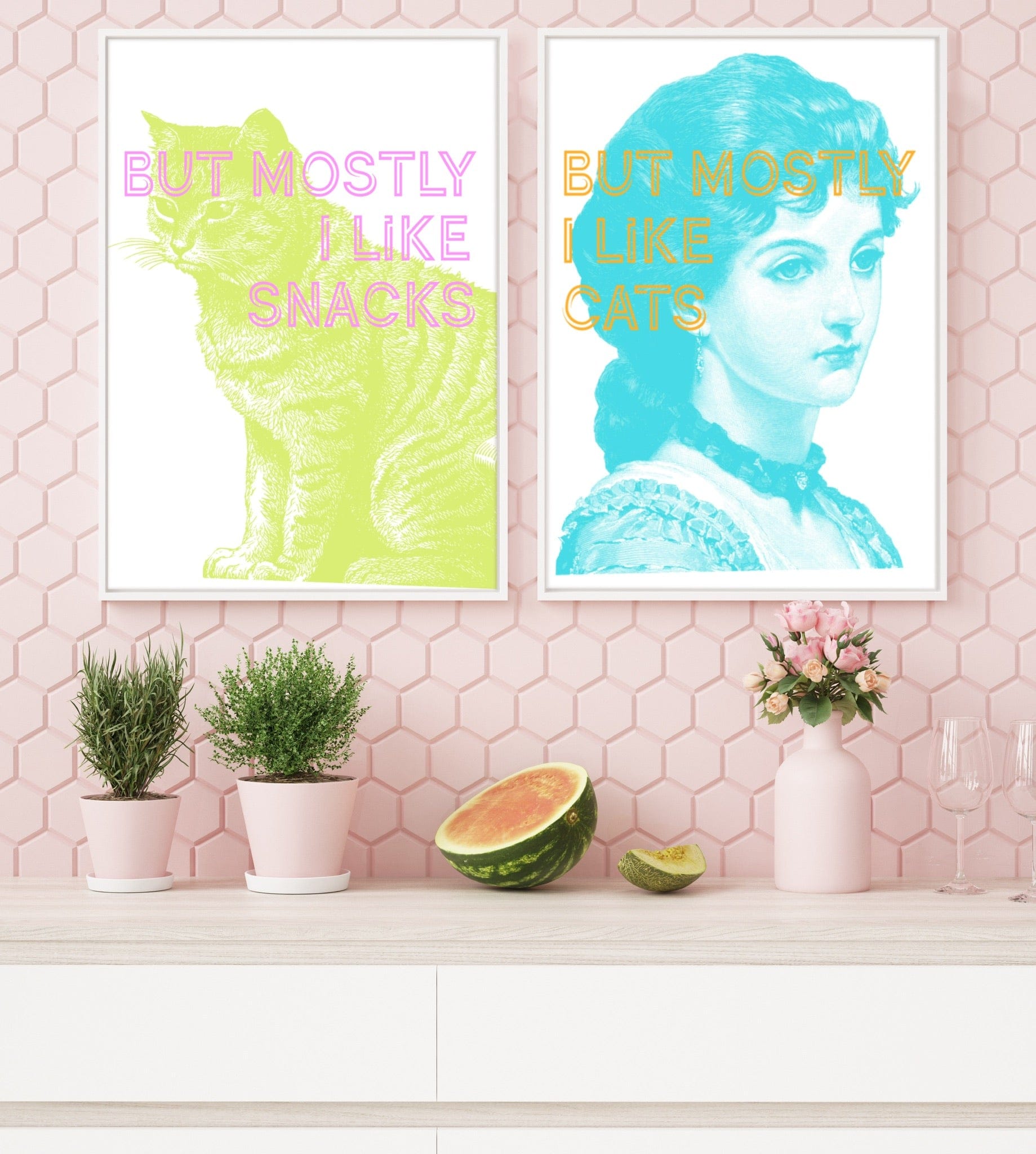 Two “But Mostly…” framed prints hang on a pink hexagon-tiled wall: the “But Mostly I Like Snacks Matte Art Print” from But Mostly… and a matching cat-themed piece. On the counter are plants, fruit, and glasses.