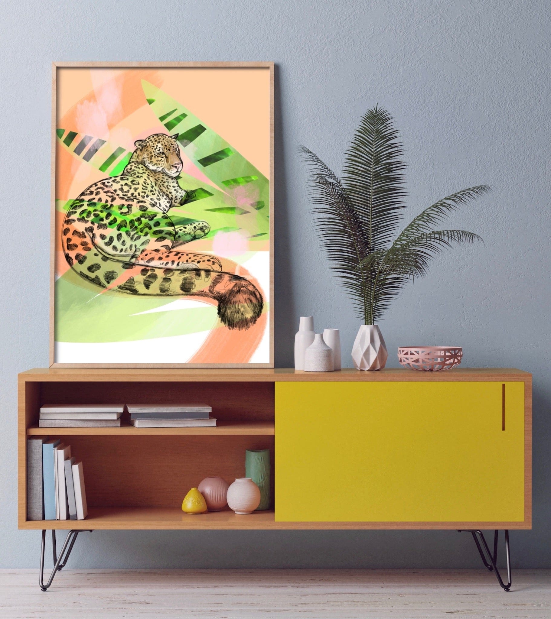 The Lounge Leopard Giclée Art Print by Leovely Leopards is displayed on a wooden sideboard with a yellow drawer, surrounded by vases, a plant, and a bowl against a gray wall—a striking tribute to big cats.