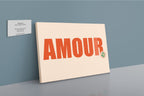 The Shromps Amour Matte Canvas Print features "AMOUR" in bold orange letters and is displayed against a blue wall next to a gallery plaque detailing artist information.