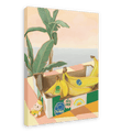 A vibrant Chonkita Bananas Giclée Canvas Print by Hôtel Dodo features bananas nestled in a cardboard box atop a checkered surface, accompanied by a small teddy bear, lush tropical plant, and the ocean beyond—capturing the whimsical charm of Hôtel Dodo, where even a banana cat may stop by.
