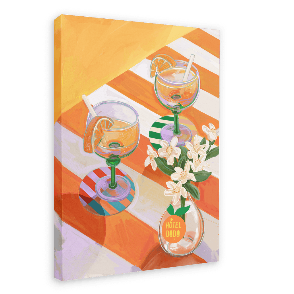 A L’Orange Giclée Canvas Print by Hôtel Dodo depicts two cocktails with orange slices and straws on a striped table, complemented by curated table settings and a small vase of white flowers marked with the brand’s name.