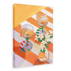 A L’Orange Giclée Canvas Print by Hôtel Dodo depicts two cocktails with orange slices and straws on a striped table, complemented by curated table settings and a small vase of white flowers marked with the brand’s name.
