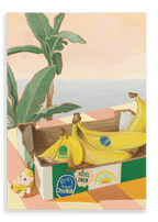 A Chonkita Bananas Giclée Canvas Print by Hôtel Dodo—featuring a lovable banana cat cozily nestled in its peel—rests on a checkered table beside a plant, with an assortment of bananas adorned with colorful stickers and a small figurine lounging nearby.