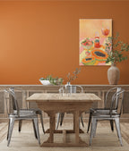 A wooden dining table with four metal chairs sits in front of an orange wall, adorned with Hôtel Dodo’s L’Aperitivo Leopardo Giclée Canvas Print and a large vase with branches—evoking the relaxed charm of an Indian summer picnic at Hôtel Dodo.