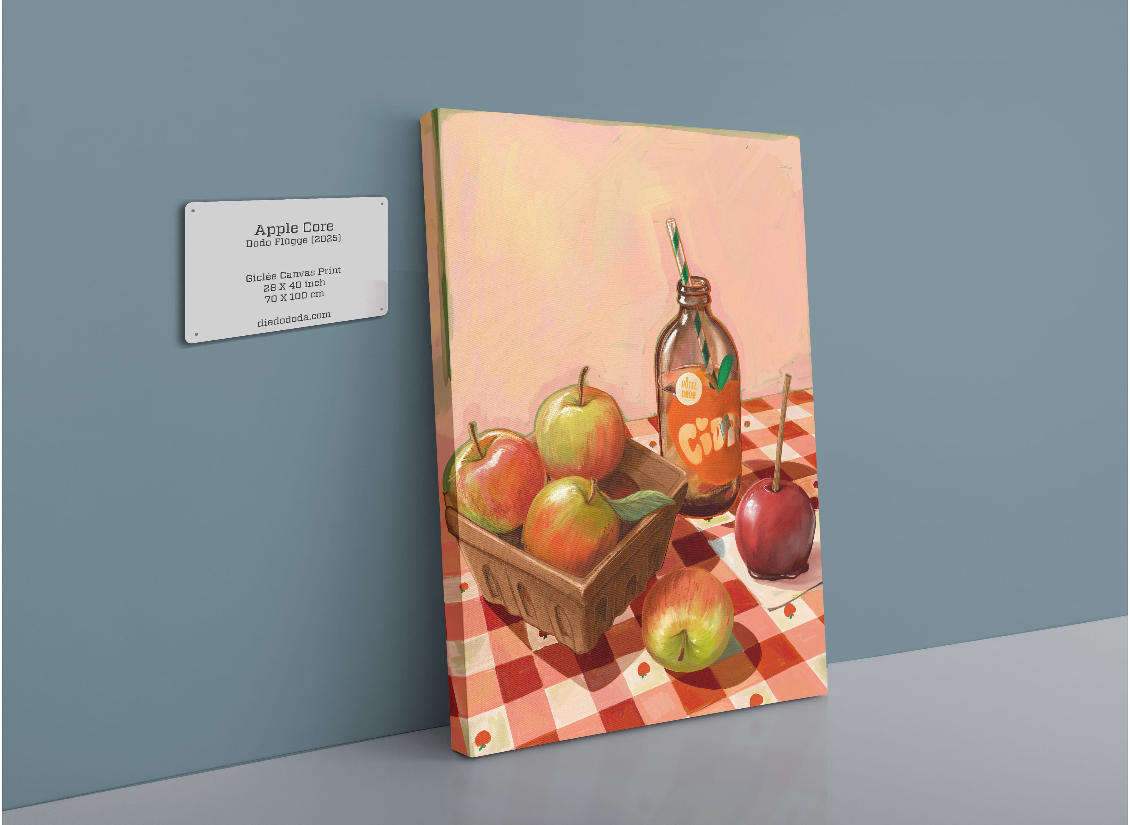 A canvas print by Hôtel Dodo, titled “Apple Core Giclée Canvas Print,” depicts apples, a basket, and a glass bottle resting on a checkered tablecloth—evoking the charm of an apple orchard in autumn. A wall plaque beside it displays the product name and brand.