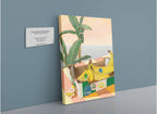 A Chonkita Bananas Giclée Canvas Print by Hôtel Dodo rests on a ledge near a potted plant with an ocean view at Hôtel Dodo; a small toy and three banana cat stickers are also visible on the ledge.