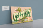 The Shromps Vintage Greetings Matte Canvas Print, featuring large “GREETINGS” text with palm trees and postcard-style stickers, is displayed on a shelf beside a business card against a light blue wall.