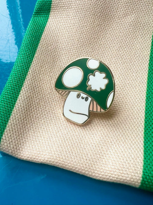 The Irish Luckshroom Pin by Pins by diedododa features a green mushroom with a neutral expression on beige fabric with green stripes, set against a blue background—a whimsical addition to any collection.