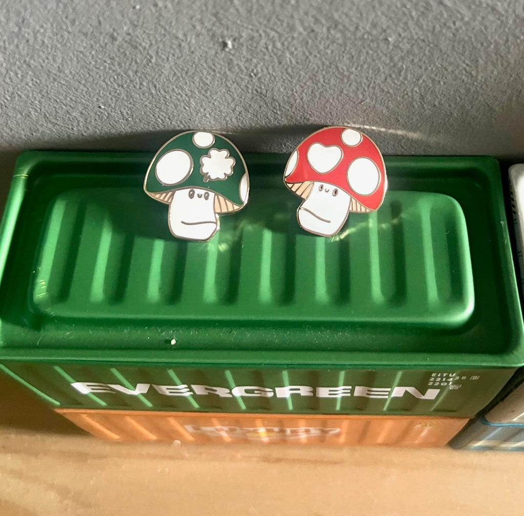 The Irish Luckshroom Pin set by Pins by diedododa features two mushroom-shaped pins—a green one with a white flower and a red one with white hearts—displayed on a green "EVERGREEN" box. Add whimsy and luck to your look!.