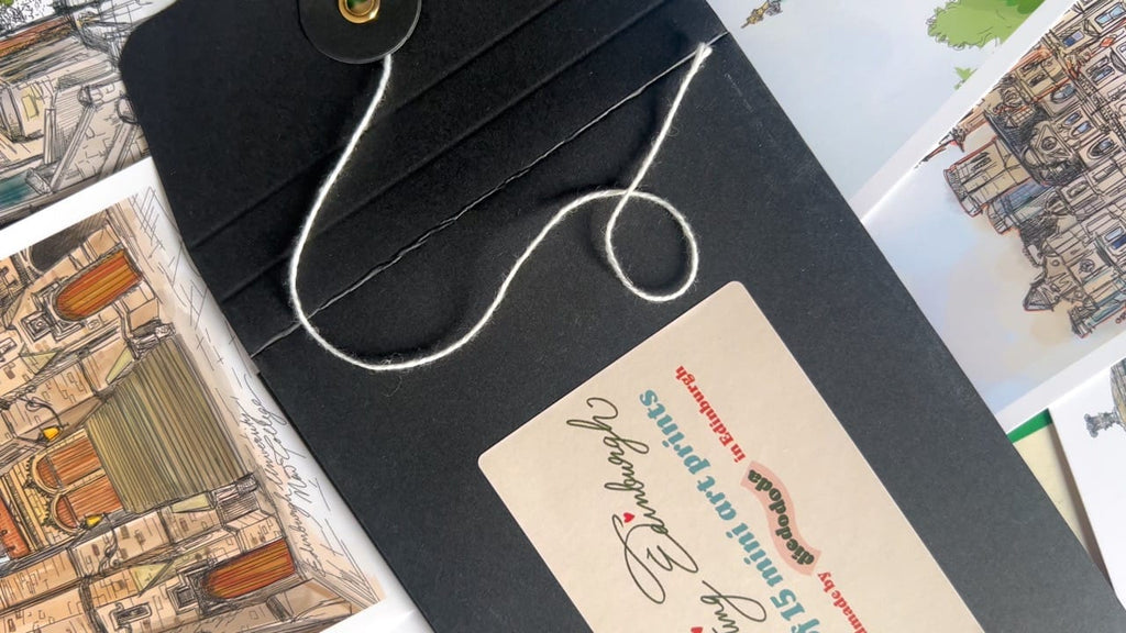 A black envelope with string closure rests on illustrated Edinburgh art prints, labeled “Edinburgh Mini Print Set | 15 A6 Original Drawings” by Essential Edinburgh—an ideal Scotland gift or unique souvenir.