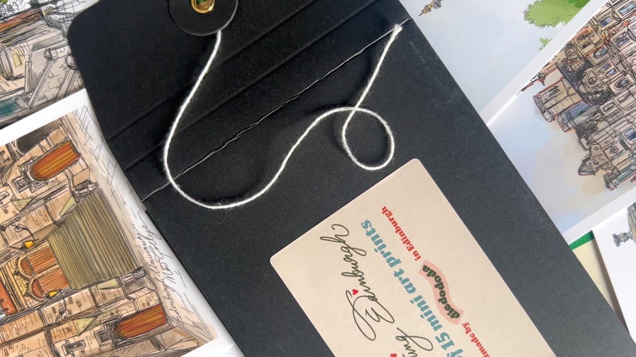 A black envelope with string closure rests on illustrated Edinburgh art prints, labeled “Edinburgh Mini Print Set | 15 A6 Original Drawings” by Essential Edinburgh—an ideal Scotland gift or unique souvenir.