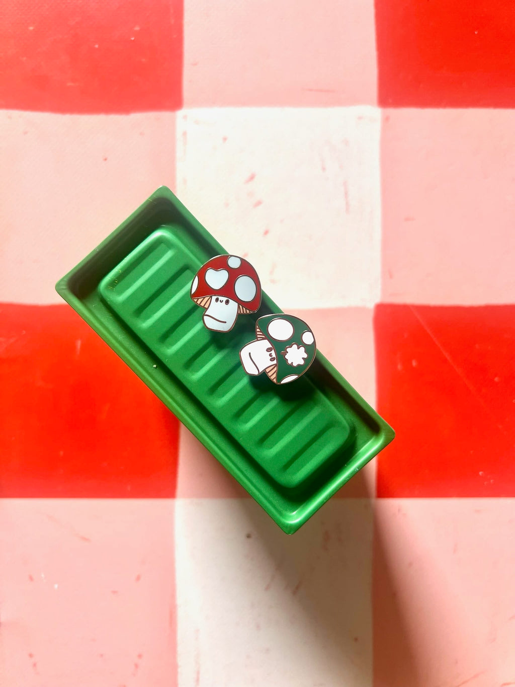 Two mushroom-shaped pins—a green mushroom pin and the Irish Luckshroom Pin by Pins by diedododa—sit on a green rectangular container, placed on a red and white checkered surface.