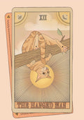 The Hanged Man Art Print by Tarot Cats features an orange cat hanging upside down from a tree branch, set against a backdrop of a sun and abstract patterns.