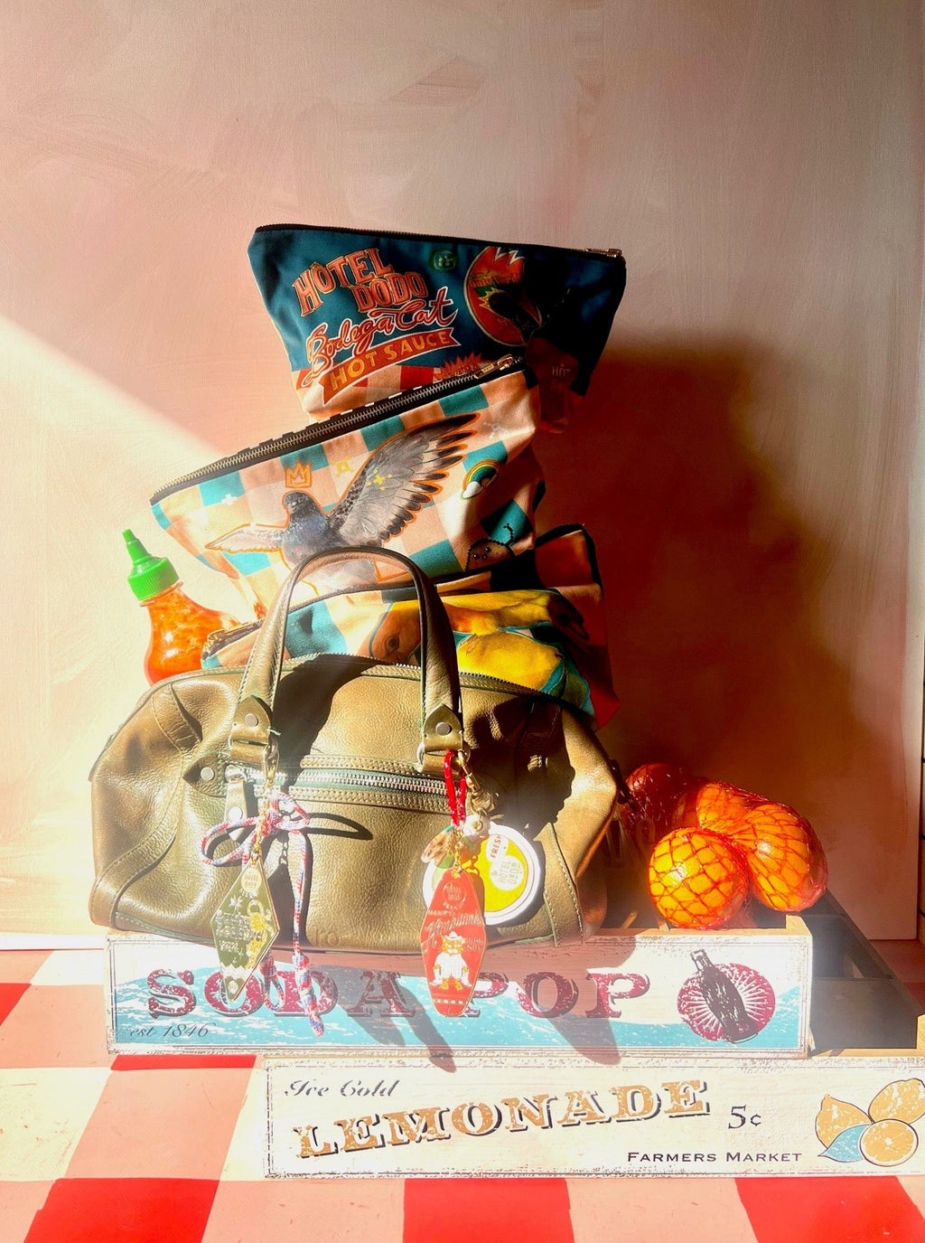 A Chaos Creatures Pouch by Aventures Des Créatures, travel bags, a leather handbag, and oranges rest on a vintage “Lemonade” crate with a checkered top in bright sunlight.