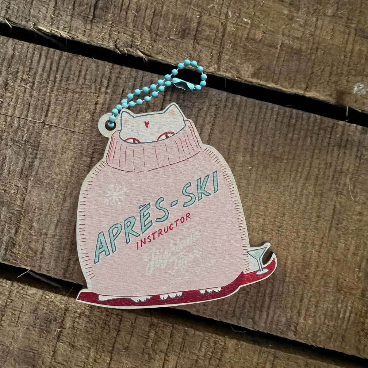 The Ski Instructor Hanging Decoration by Christmas by diedododa features a wooden tag depicting a cat in a pink sweater labeled “APRES-SKI INSTRUCTOR,” partially snuggled inside its cozy outfit. Lying on a wooden surface, this eco-friendly decoration makes a delightful addition to your collection of gift tags or wooden animal Christmas ornaments.
