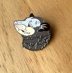 The Doing My Pest Pin by Pins by diedododa is a cute enamel pin with a cartoon opossum head and "doing my best" text on its body, shown here on a wooden surface.