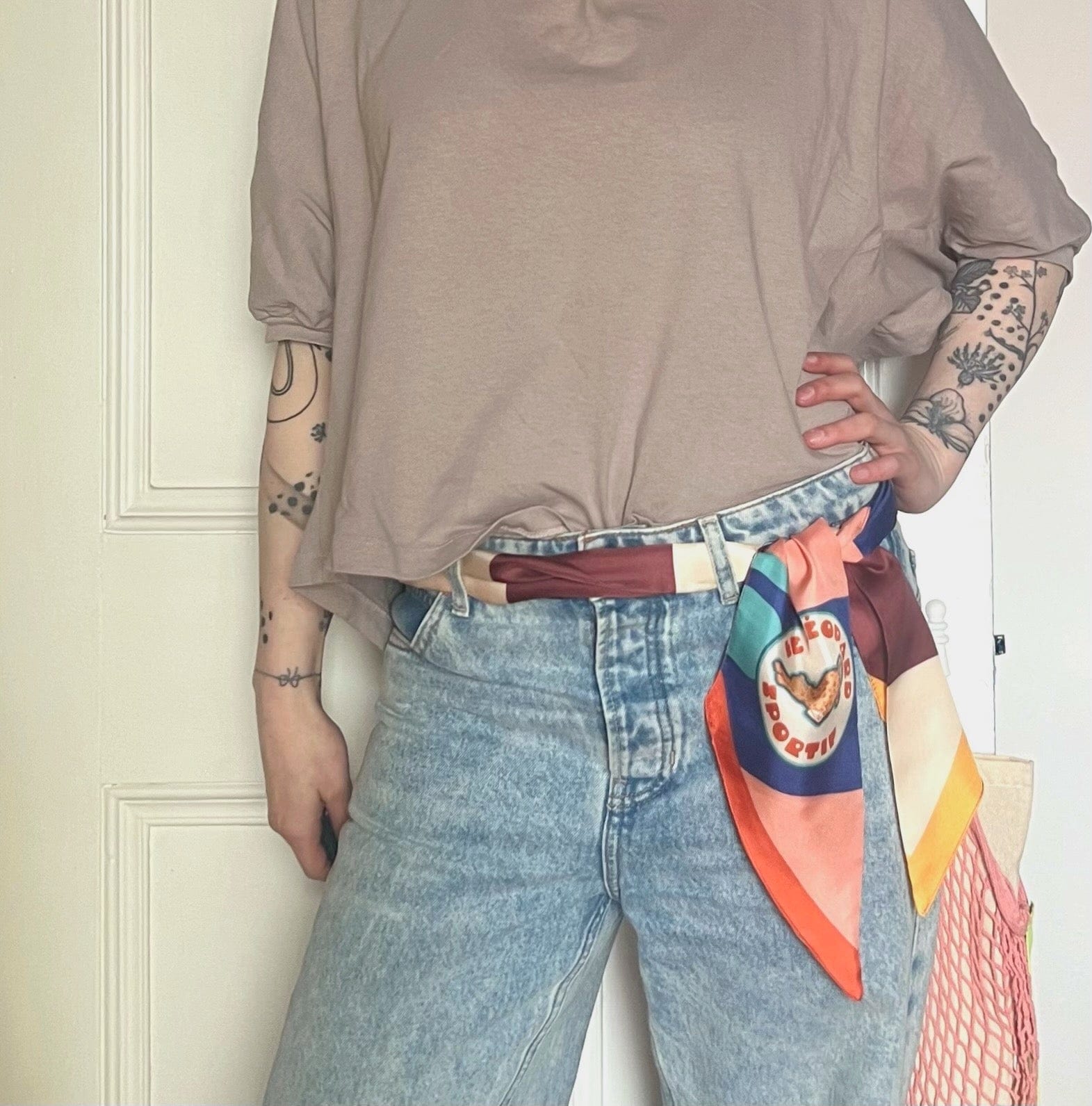 A person styled in a loose beige top, light blue jeans, and the diedododa Hotel Dodo Scarf tied around their belt loop; tattoos are visible on both arms.