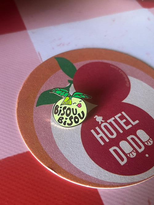 The Mello Pomelo Pin from Pins by diedododa sits on a circular mat labeled "HOTEL DODO," featuring vibrant citrus graphics, all placed on a red and white checkered surface.