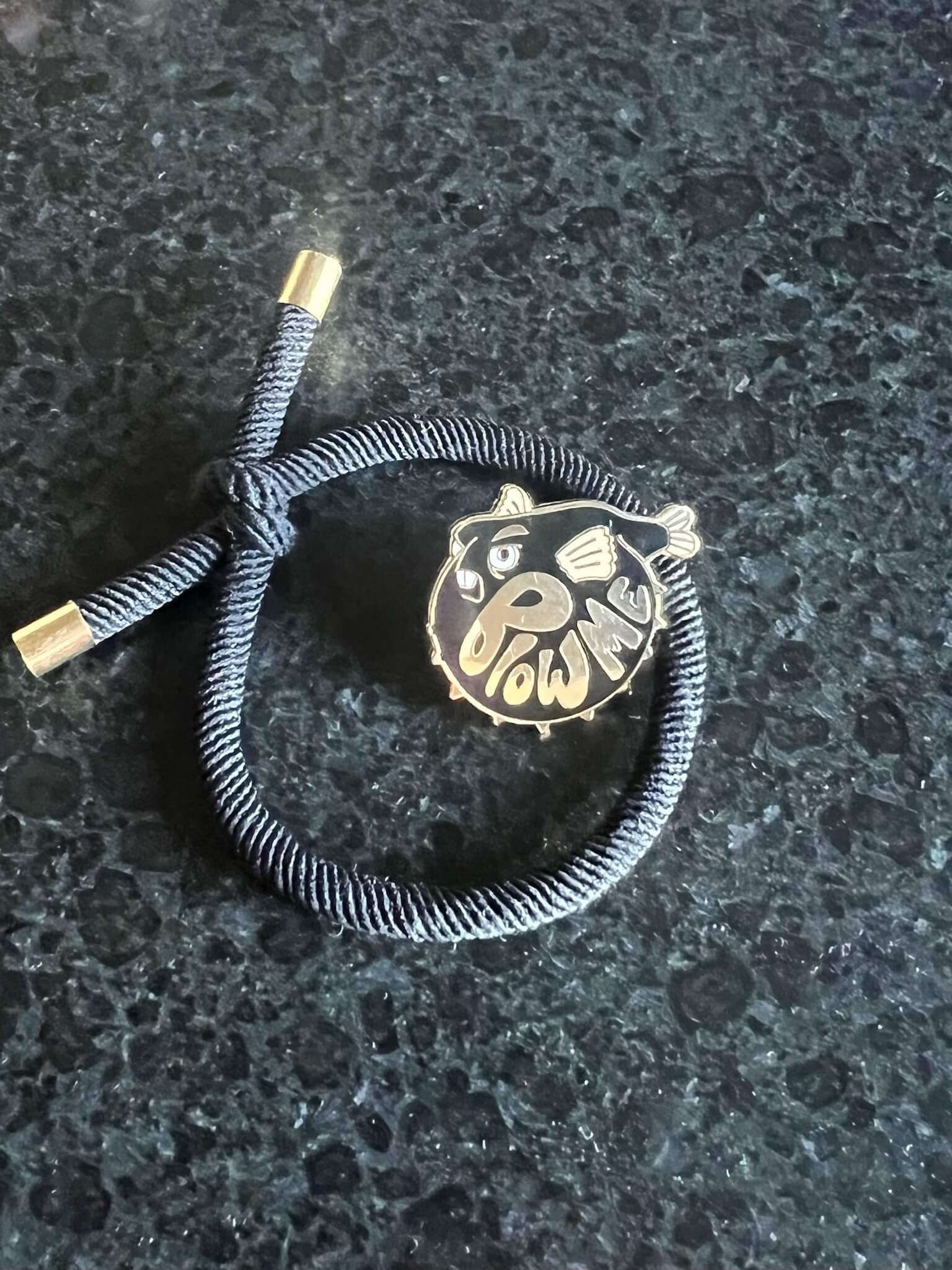 A dark bracelet sits on a deep surface, featuring a gold-accented knot and a round charm with the Pins by diedododa Beastly Blowfish cartoon, alongside the playful text 