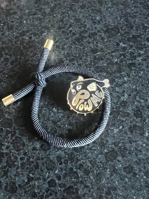 A dark bracelet sits on a deep surface, featuring a gold-accented knot and a round charm with the Pins by diedododa Beastly Blowfish cartoon, alongside the playful text "WOW ME.