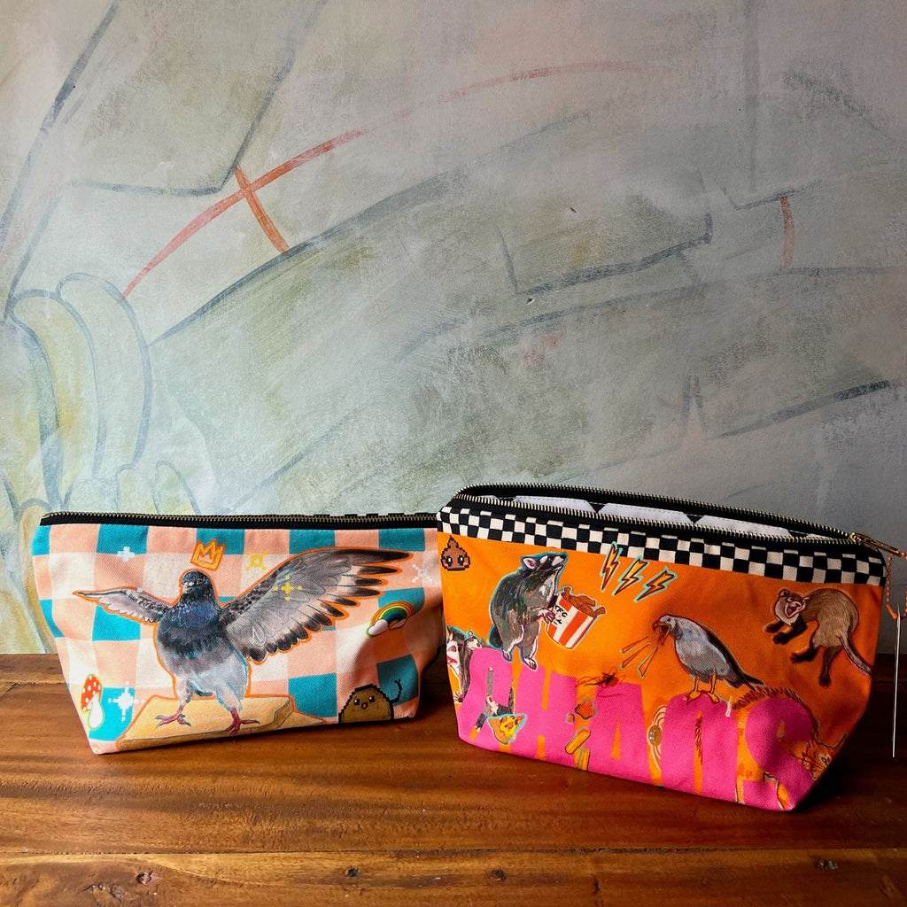 Two Chaos Creatures Pouches by Aventures Des Créatures, handmade in the UK, feature colorful birds, cats, and geometric designs. Displayed on wood with a painted wall backdrop.