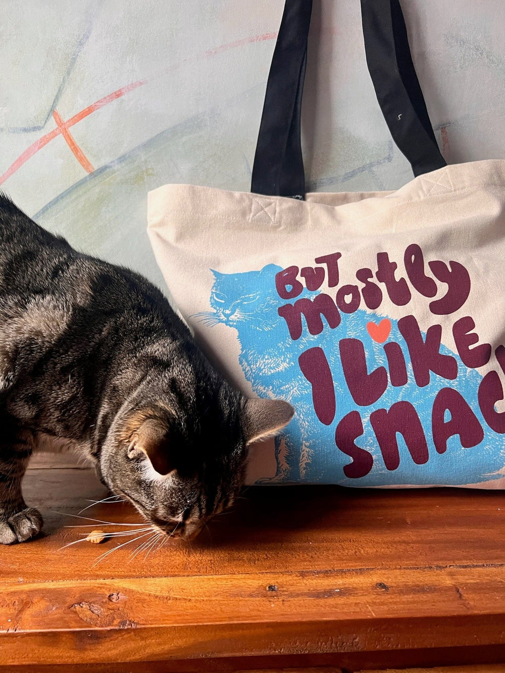 A gray tabby cat curiously sniffs a wooden surface beside a cotton canvas bag inscribed with "But Mostly I Like Cats." The shopping and beach bag from Bags by diedododa hangs gracefully against a light-colored wall, adding charm to the scene.