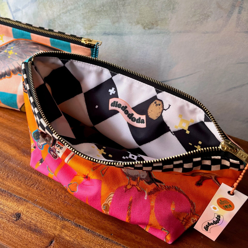 A zippered cotton Chaos Creatures Pouch by Aventures Des Créatures, with a colorful animal design and checkered lining, sits open on a wooden surface showing its label. Handmade in the UK. Another pouch is partially visible behind it.