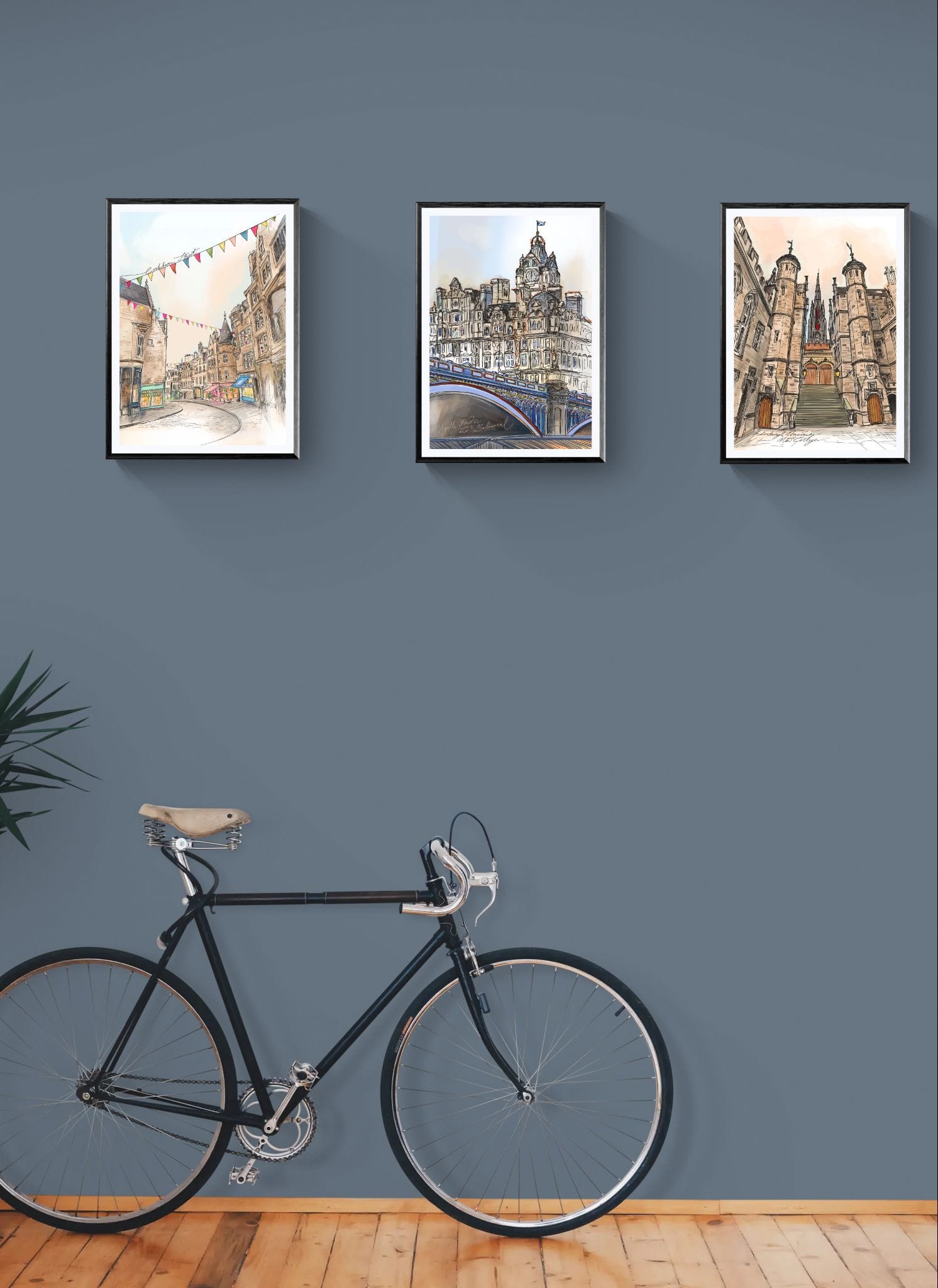 A black bicycle rests against a blue wall beneath three framed "North Bridge & The Balmoral Edinburgh Matte Art Prints" by Essential Edinburgh, with a plant partially visible on the left, suggesting a freshly updated space.