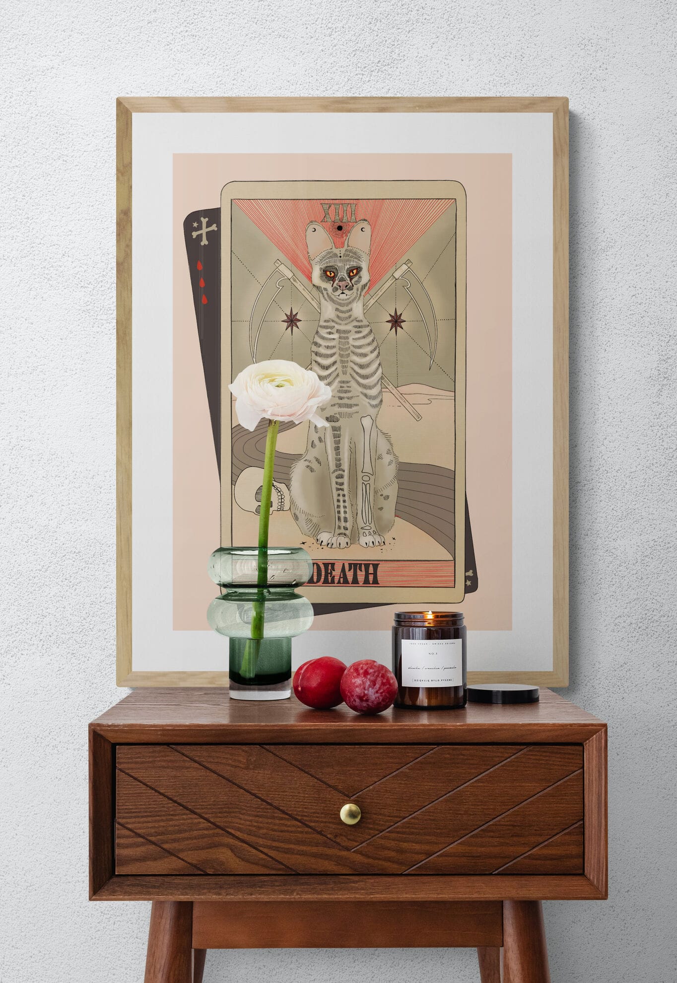 The Death Matte Art Print by Tarot Cats, inspired by vintage tarot cards, features a skeletal cat as the Death card. It's shown on a wall above a wooden table with a flower vase, two plums, and a lit candle.