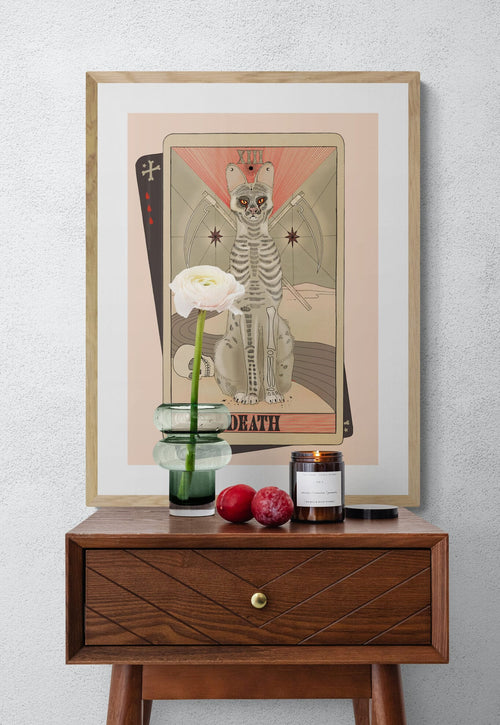 The Death Matte Art Print by Tarot Cats, inspired by vintage tarot cards, features a skeletal cat as the Death card. It's shown on a wall above a wooden table with a flower vase, two plums, and a lit candle.
