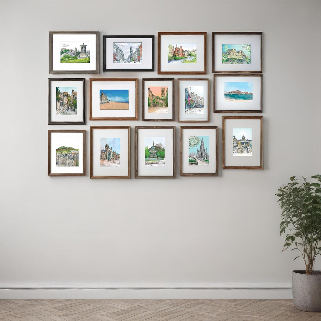 A grid of framed landscape and architectural artworks, including the Edinburgh Mini Print Set (15 A6 original drawings) by Essential Edinburgh, is displayed on a light wall above a plant in a gray pot on a wooden floor.