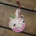 The Feral Fest Hanging Decoration by Christmas by diedododa showcases an illustrated opossum wearing a crown and holding a pickle, with the phrase “Have a Feral Fest.” Displayed on a wooden surface, it’s the perfect festive accent to pair with eco-friendly gift tags or wooden animal Christmas decorations during your celebrations.
