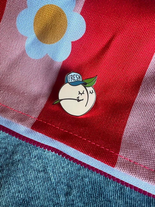 This charming spring accessory, the Farm Fresh Peach Pin by Pins by diedododa, showcases a cartoon peach with a green leaf and "Fresh" hat, beautifully paired with red and pink striped fabric featuring an uplifting daisy.