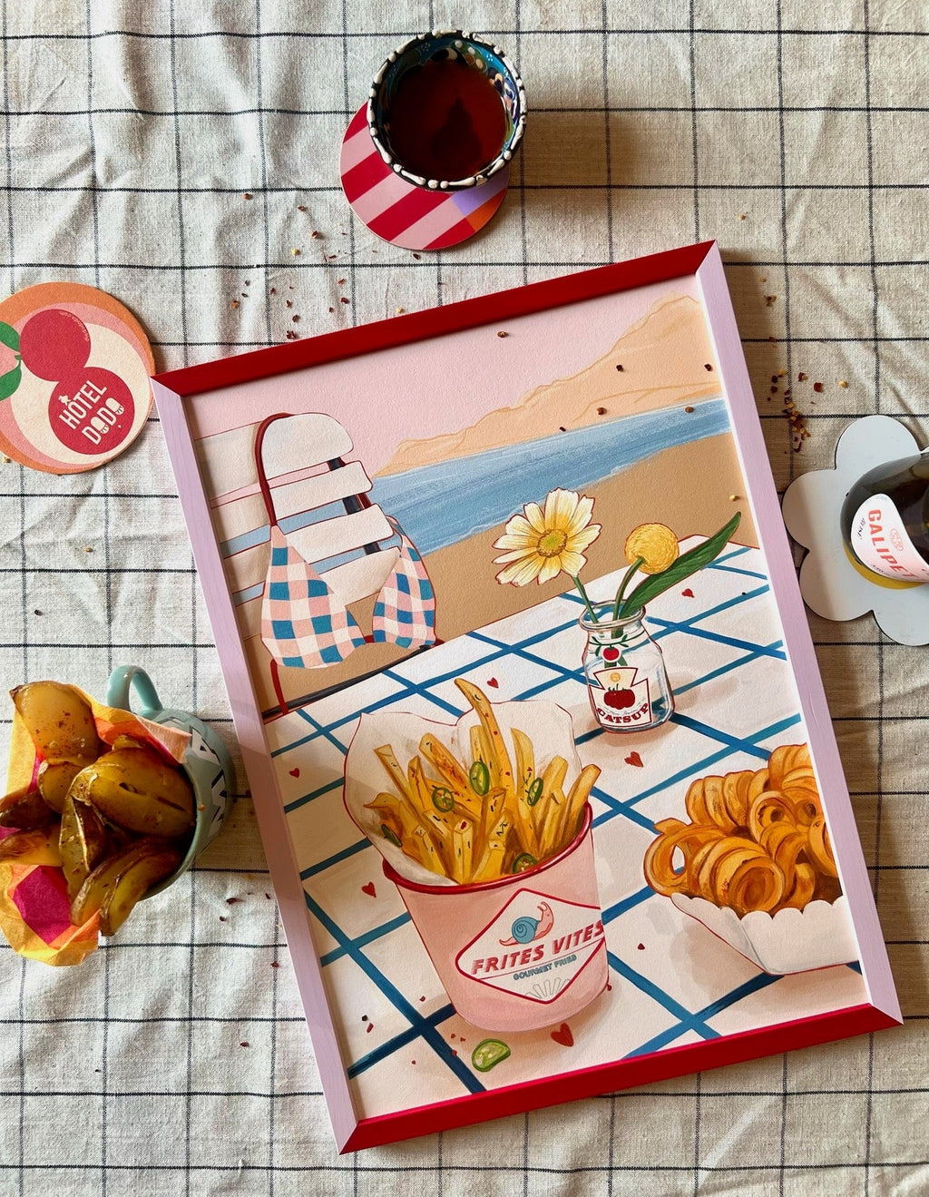 The Frites Vites Giclée Art Print by Hôtel Dodo features gourmet fries, curly fries, and drinks on a tiled table by the sea, beautifully displayed on a checkered cloth with snacks and coasters, overlooking a stunning ocean view.