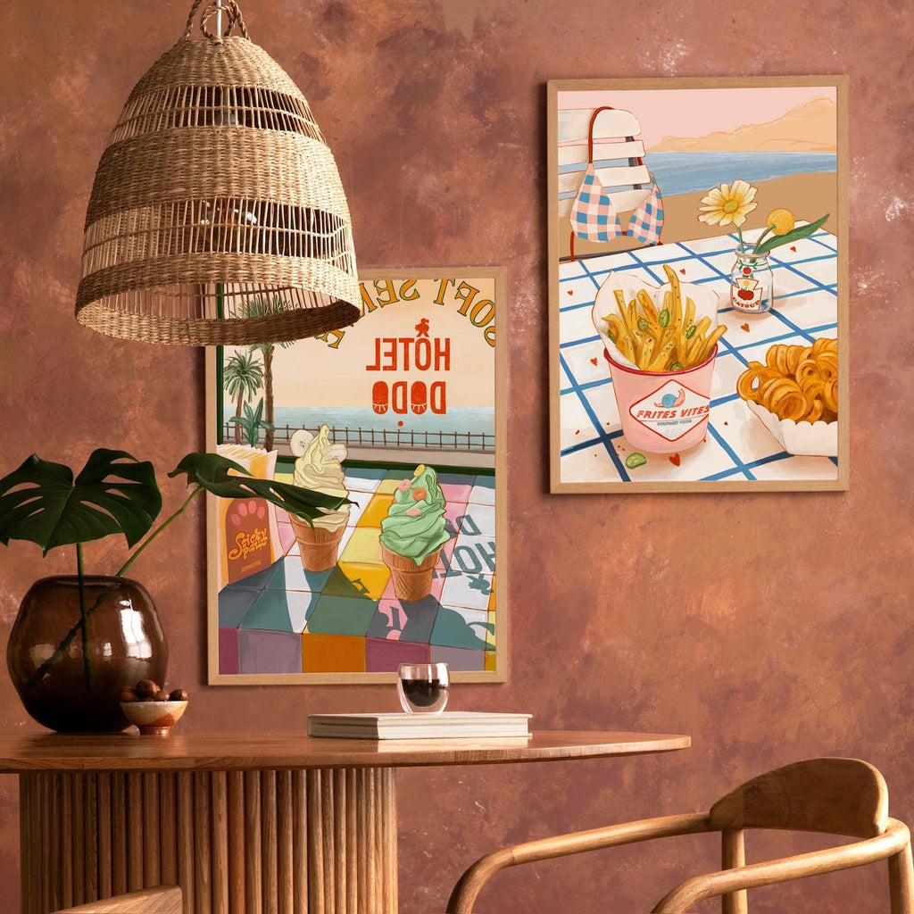 A room with a round wooden table, a plant, a rattan pendant lamp, and two framed posters—one being the Hôtel Dodo "Frites Vites" Giclée Art Print—displaying summery food illustrations on a textured brown wall.