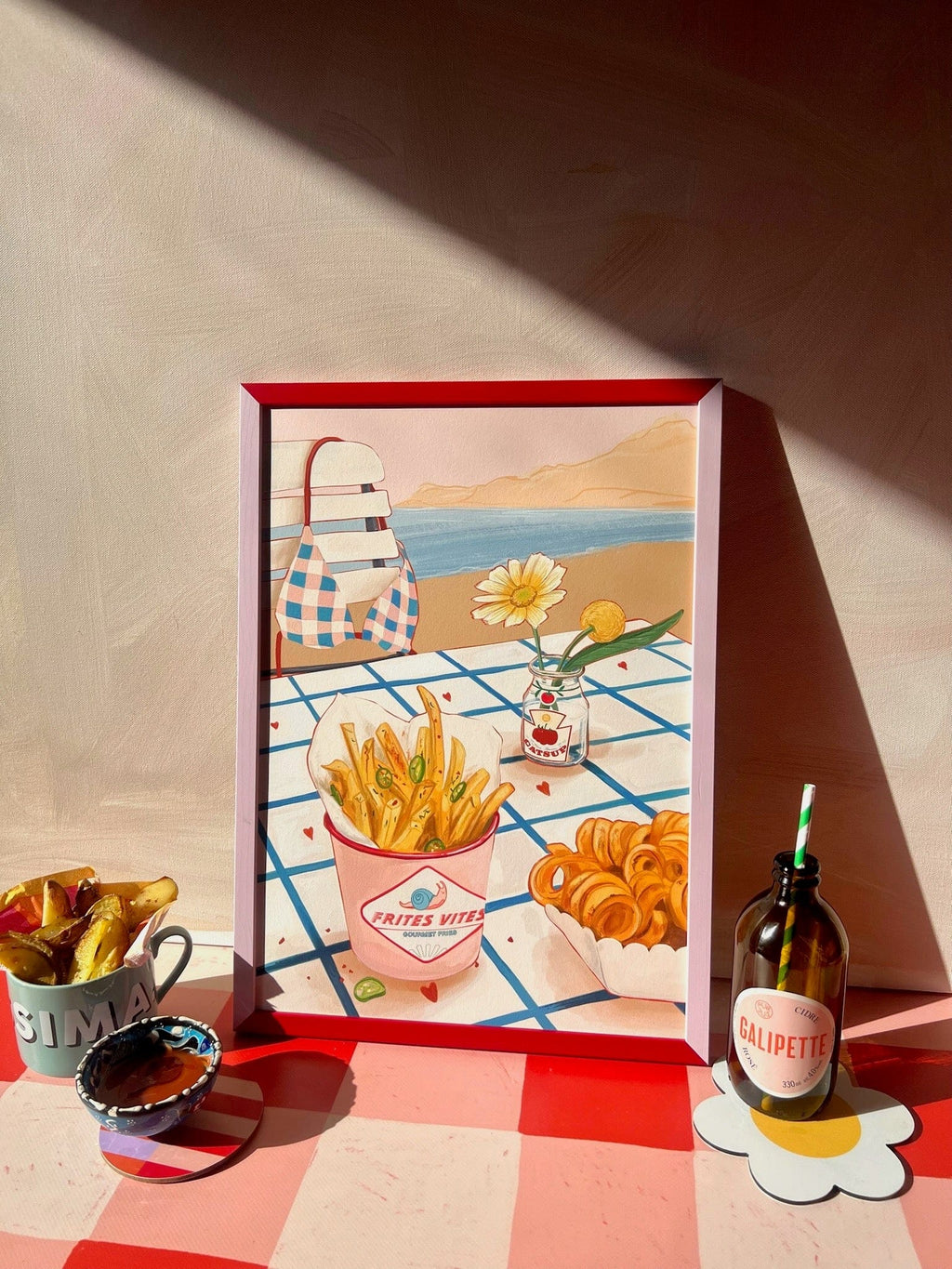 The Frites Vites Giclée Art Print by Hôtel Dodo features gourmet fries and drinks on a checkered table, shown with a mug of fries, a small flower jar, and a bottle with a straw on the same checkered surface.