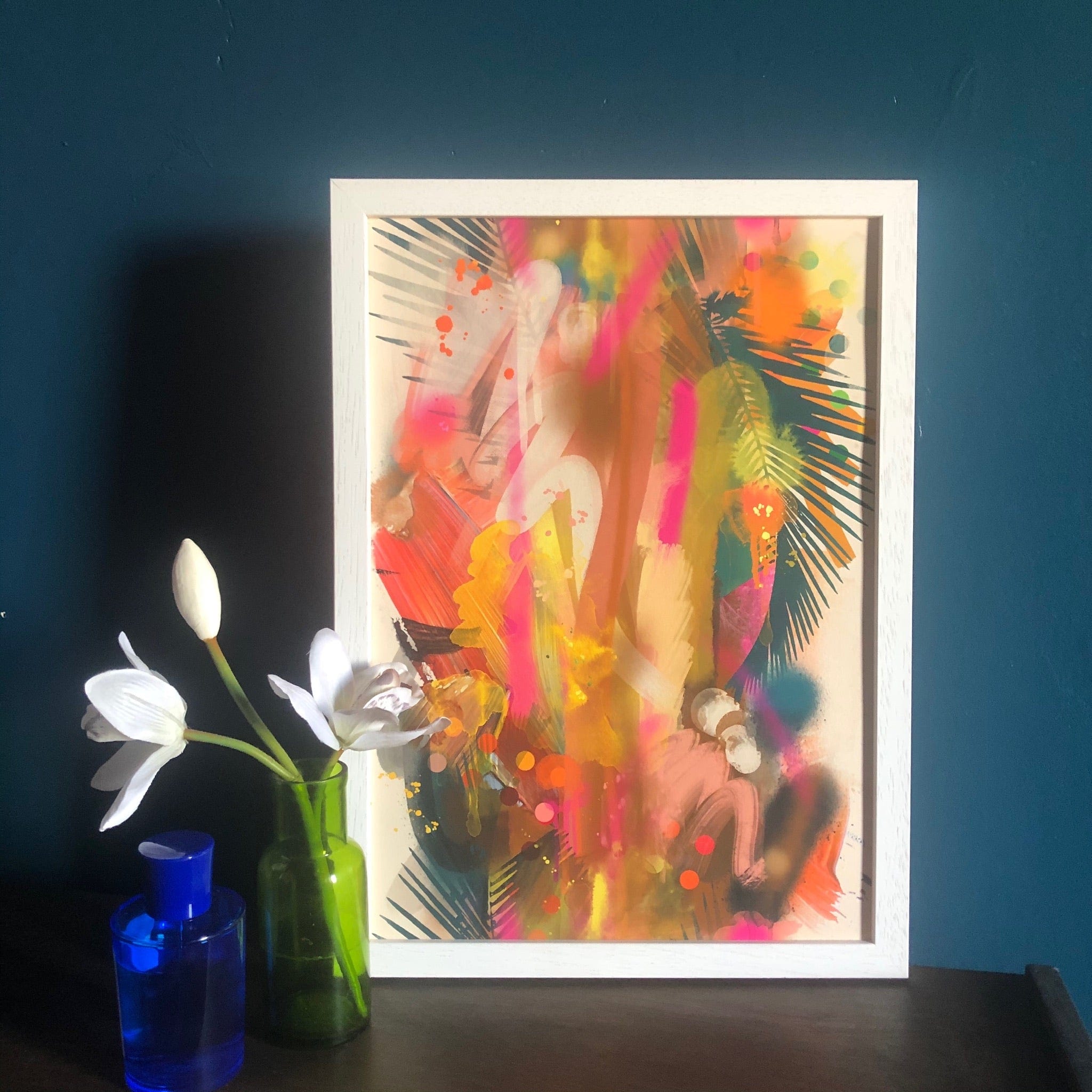 The Miami Vice Giclée Art Print by Road Tripping, with its vibrant abstract colors, stands beside a green vase of white flowers and blue bottle on a dark surface, capturing the essence of Miami escapes against a dark teal wall.