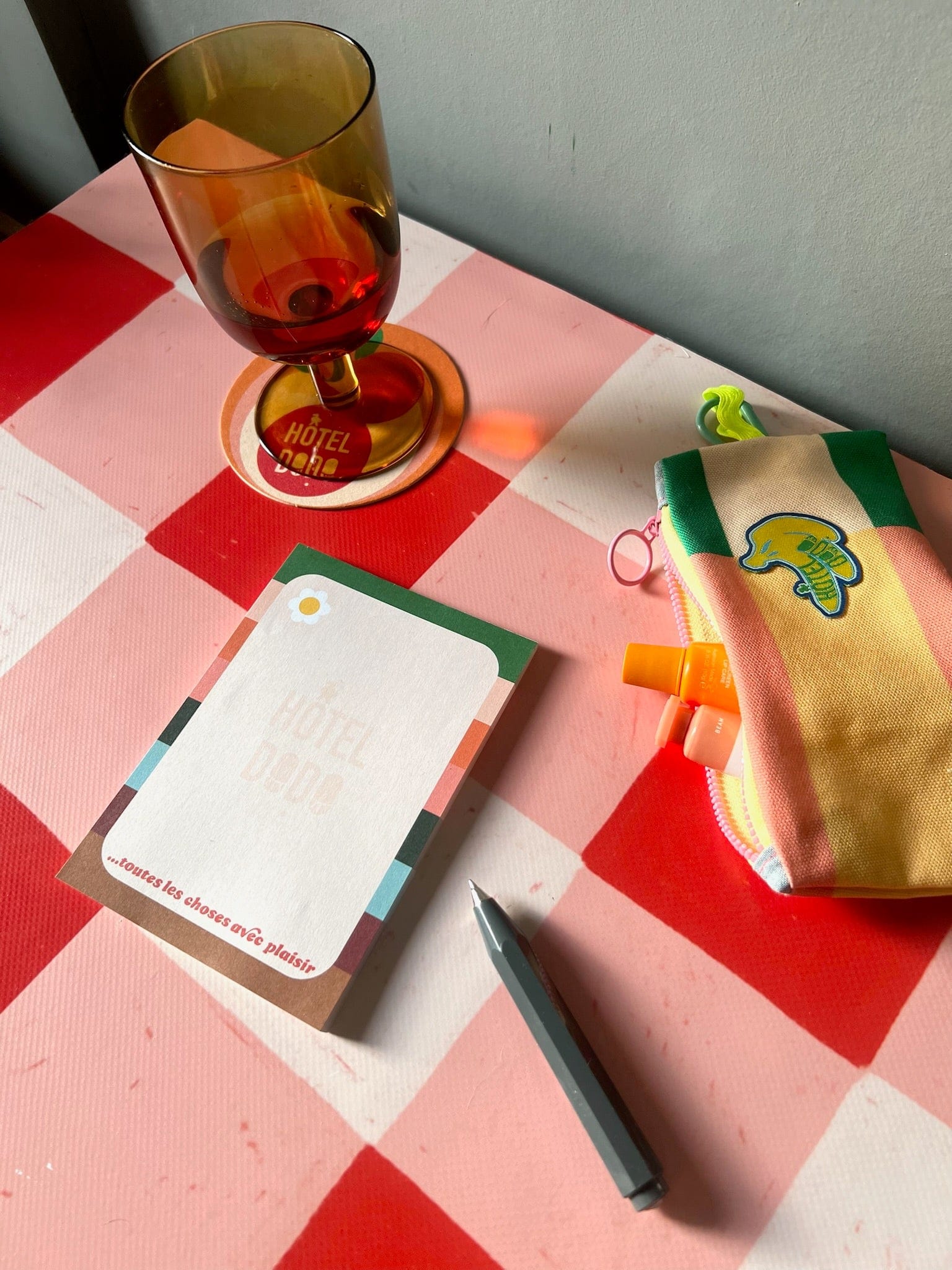 A glass with a drink, hotel stationery, Hôtel Dodo “All The Things With Pleasure” Hotel Style Notepad, pen, and pastel pencil pouch sit on a red and white checkered table with a touch of retro stripe charm.