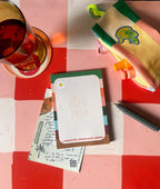 The “All The Things With Pleasure” Hotel Style Notepad by Hôtel Dodo is displayed alongside stationery, a postcard, pen, pouch, lip balm, and a glass of red liquid on a red and white checkered surface with retro stripes.