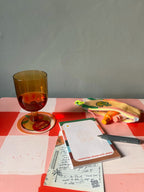 A yellow-tinted glass sits on a red checkered table beside a zippered pouch, pen, an Hôtel Dodo "All The Things With Pleasure” hotel-style notepad, and postcard under the sunny sky.