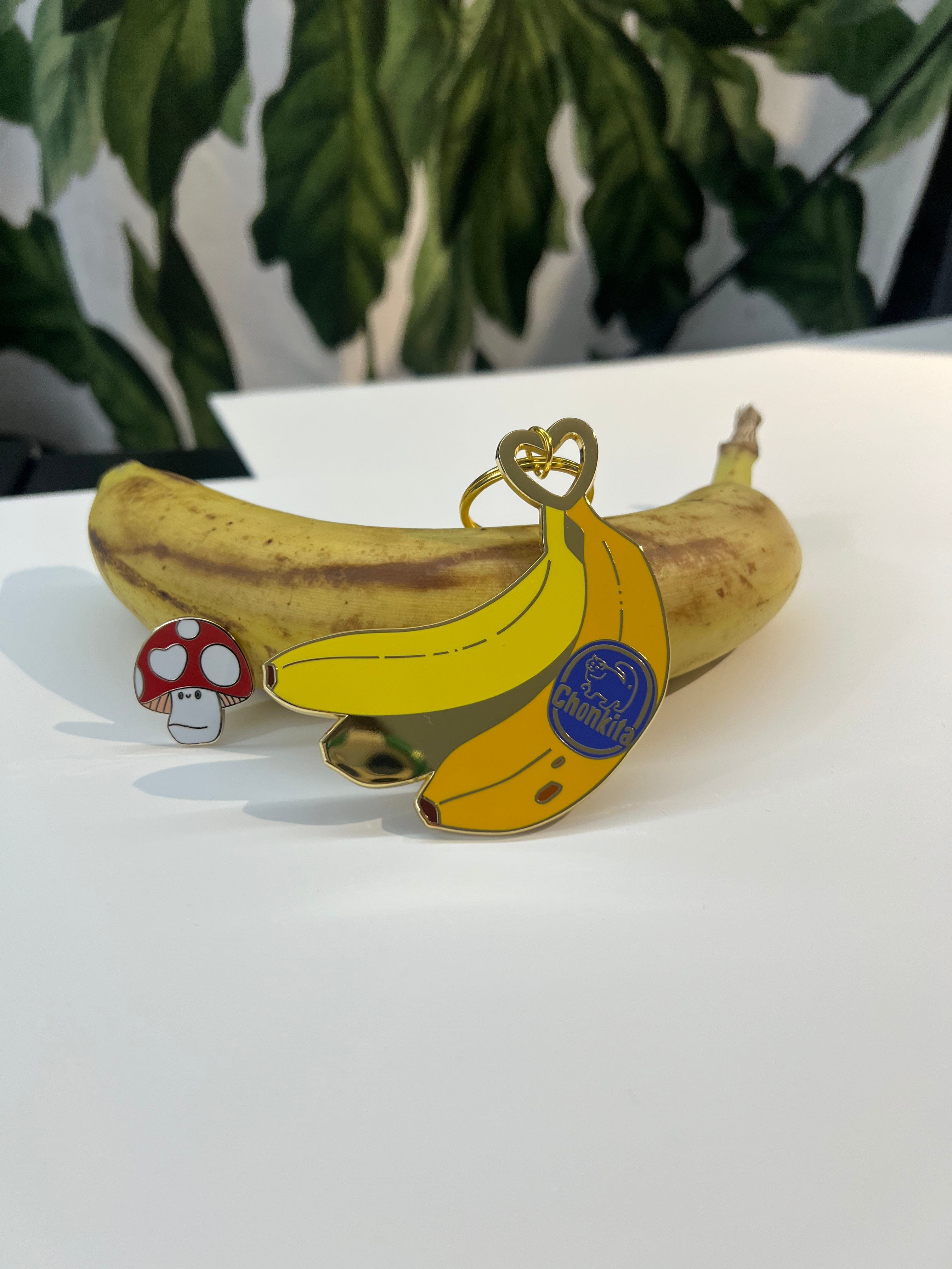 A ripe banana rests on a white surface with a cartoon banana pin and the Luckshroom Pin by Pins by diedododa—modeled after the good-luck Amanita Muscaria mushroom. Large green leaves fill the background.