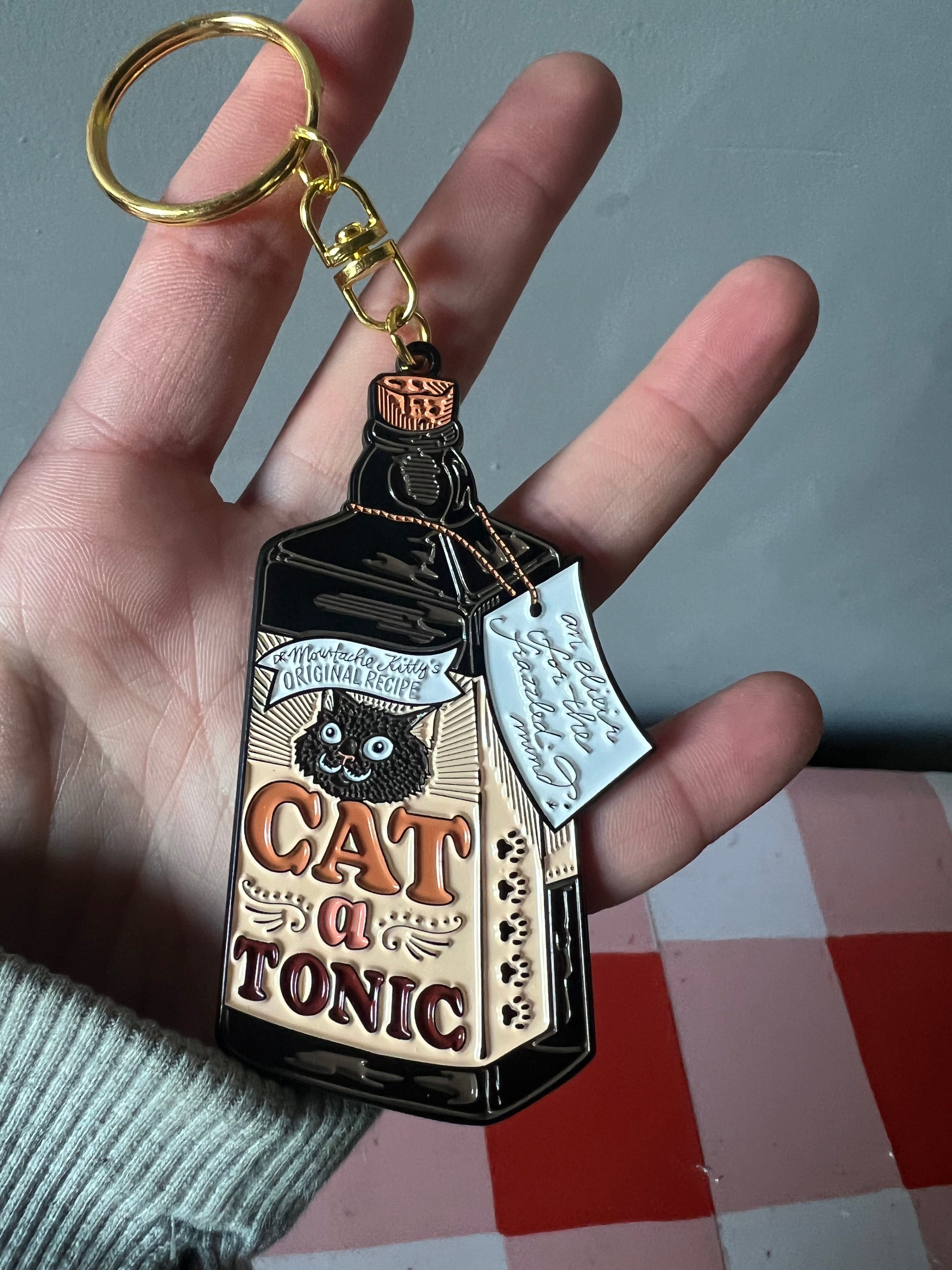 A hand holds a Catatonic Keyring by Hôtel Dodo, shaped like a vintage bottle labeled "Cat a Tonic" with a cat illustration and an attached tag.