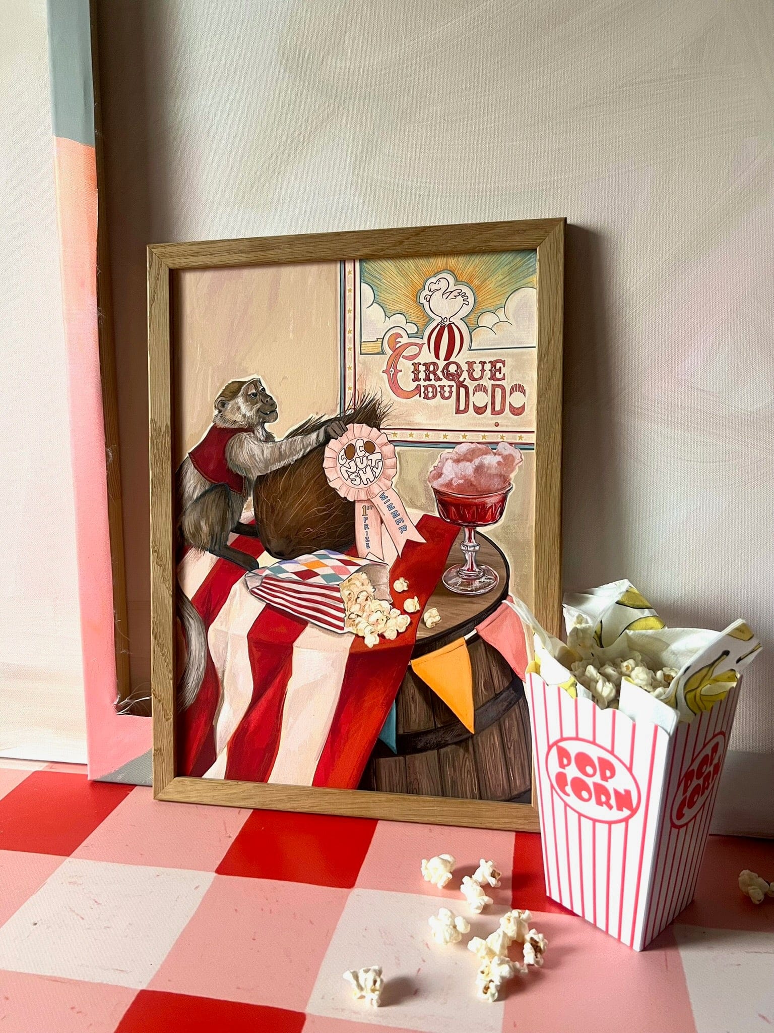 The Coconut Shy Cirque Du Dodo Giclée Art Print by Hôtel Dodo features a vintage-style monkey with circus items and popcorn on a striped tablecloth, with a box of popcorn in the foreground, recalling classic funfair charm.